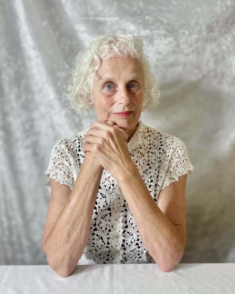buste de belle femme d'une soixantaine d'année coiffée d'un carré ondulé blanc court assise pose face à la caméra devant un fond en velour blanc.